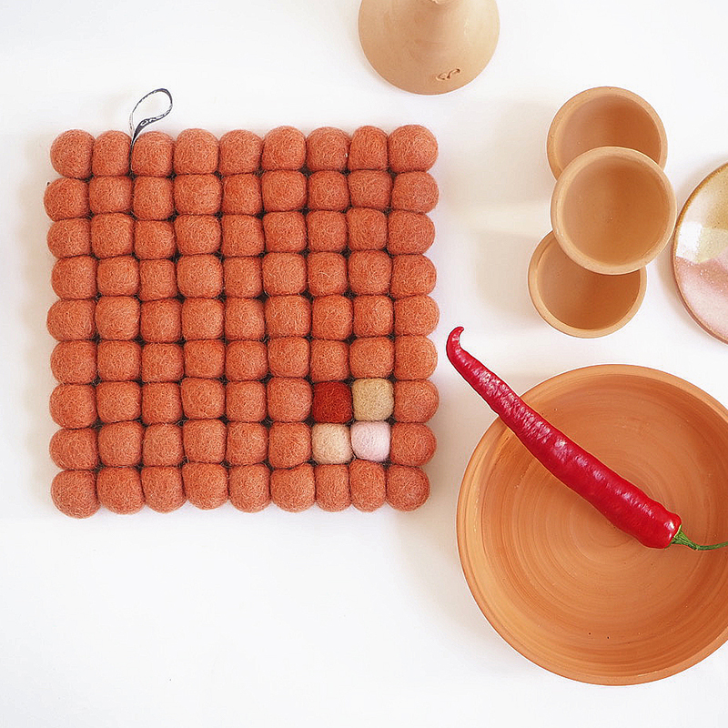 Square trivet in terracotta color made of 100% wool  with a chili and terracotta plate on the side