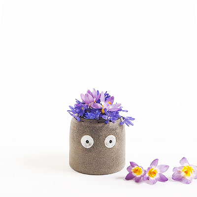Grå vas i keramik med ögon, dekorerad med lila blommor.