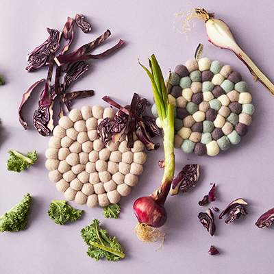 Round trivets in wool in the colors lavender, white laying on a table with vegetables.