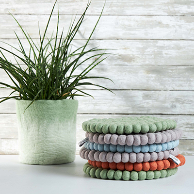 Round trivet in 100% wool in lavender, blue, green and rust in a pile, next to a green plant.