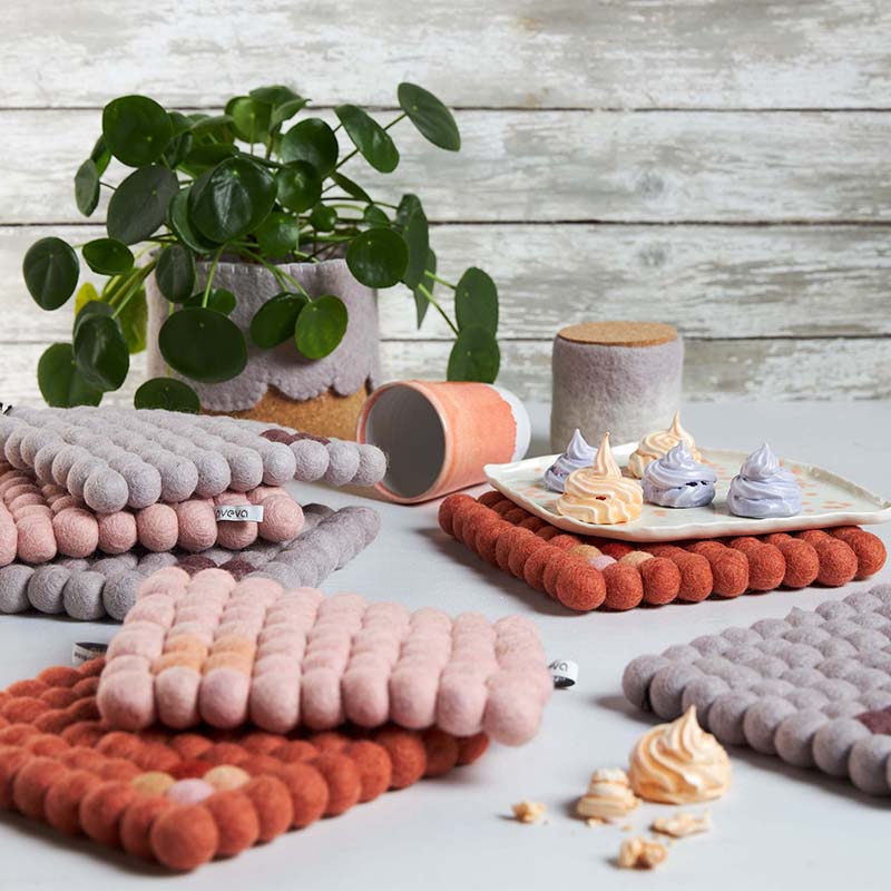Square trivet in 100% wool in lavender, pink, and rust on a table, next to a green plant.
