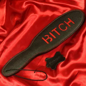 Musta nahkainen läimäyspaddle punaisella tekstillä Bitch, Chambers of Love, tuotenumerolla XB112. Täydellinen intensiivisiin ja tyylikkäisiin BDSM-sessioihin.