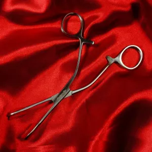 Medical clamp (pean) from Chambers of Love in high-polished stainless steel, lying on a red background that highlights its surgical precision and exclusive BDSM aesthetic.