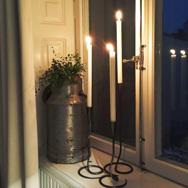 tre stycken ljusstakar i olika höjd i svart smide med tända ljus står i ett fönster på kvällen