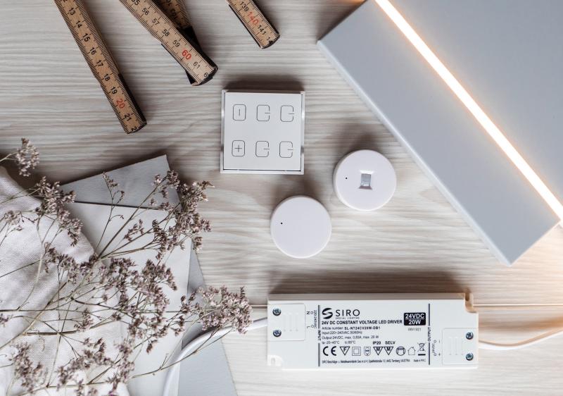 En samling belysningskomponenter, inklusive en LED-drivare, två runda sensorer och en fyrkantig kontrollpanel. Bilden inkluderar också trälinjaler och torkade blommor som dekor."