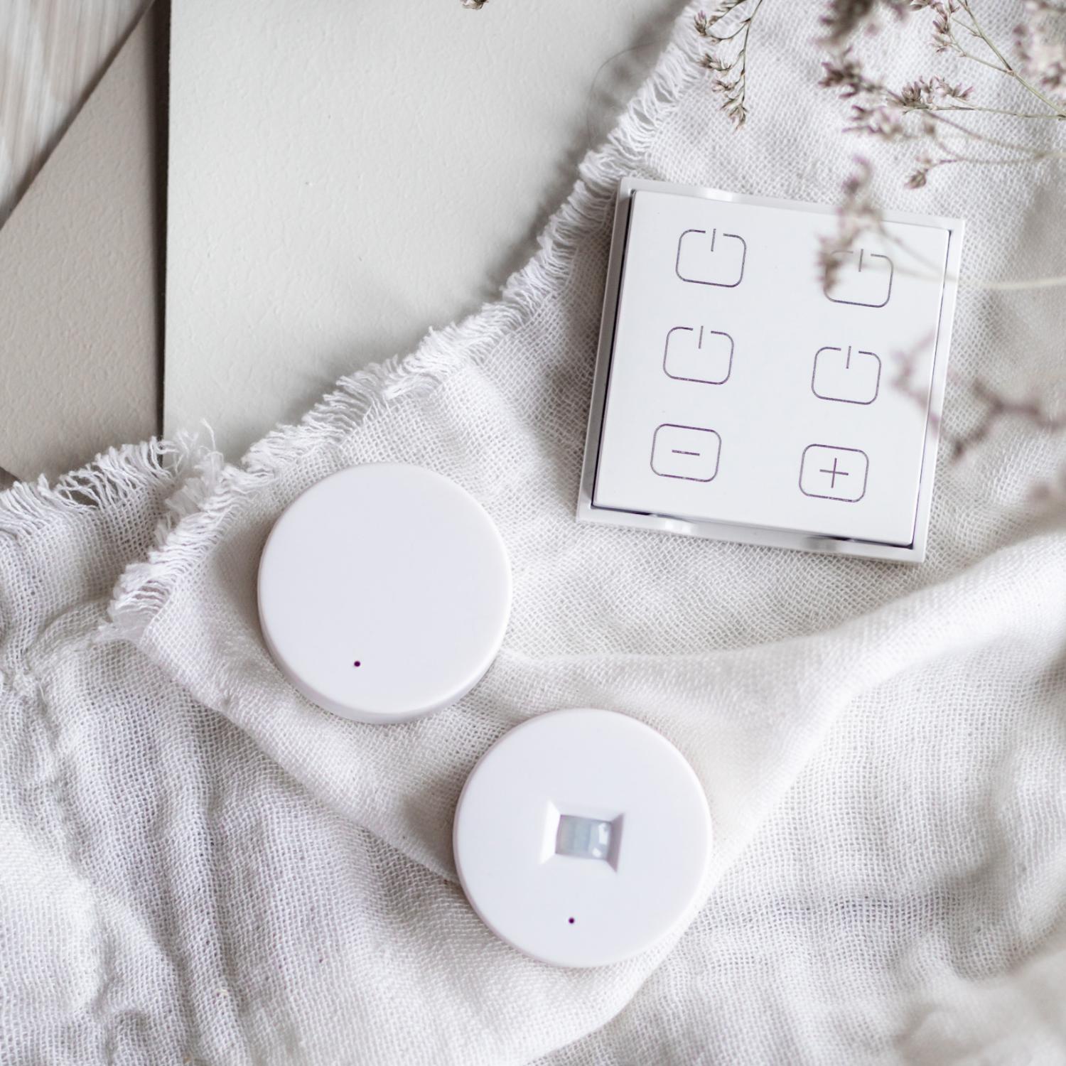 En samling belysningskomponenter, inklusive en LED-drivare, två runda sensorer och en fyrkantig kontrollpanel. Bilden inkluderar också trälinjaler och torkade blommor som dekor.