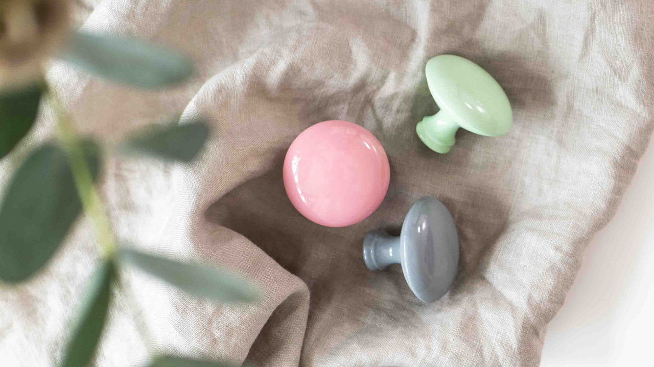 Spread joy with color in every room with knobs from the Candy Collection! 🍬 Tre färgglada knopp i pastellfärger: rosa, grå och grön, placerade på ett beige tygstycke.