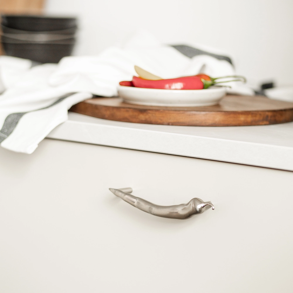 Ett silverfärgat handtag i form av en chilipeppar monterat på en vit kökslåda. I bakgrunden syns en träskärbräda med färska chilifrukter på ett litet fat.
