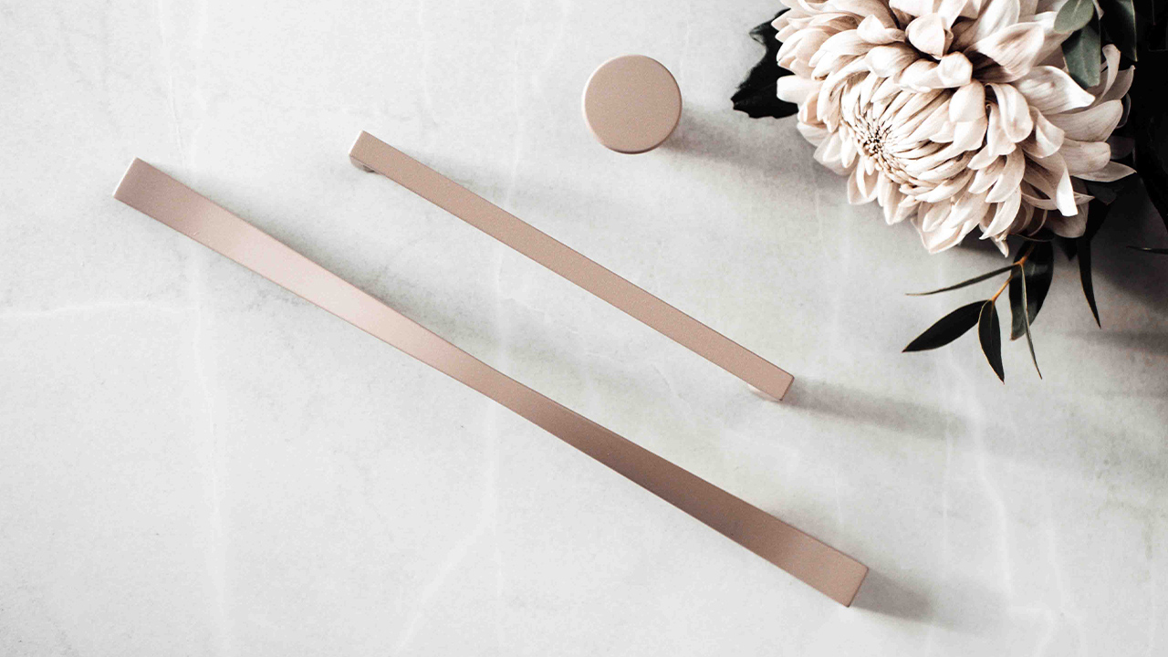 Create the right feeling with handles and knobs in delightful colors Elegant beslag i borstad metall, en rund knopp och två långa handtag, placerade på marmorbakgrund med blomdekoration.