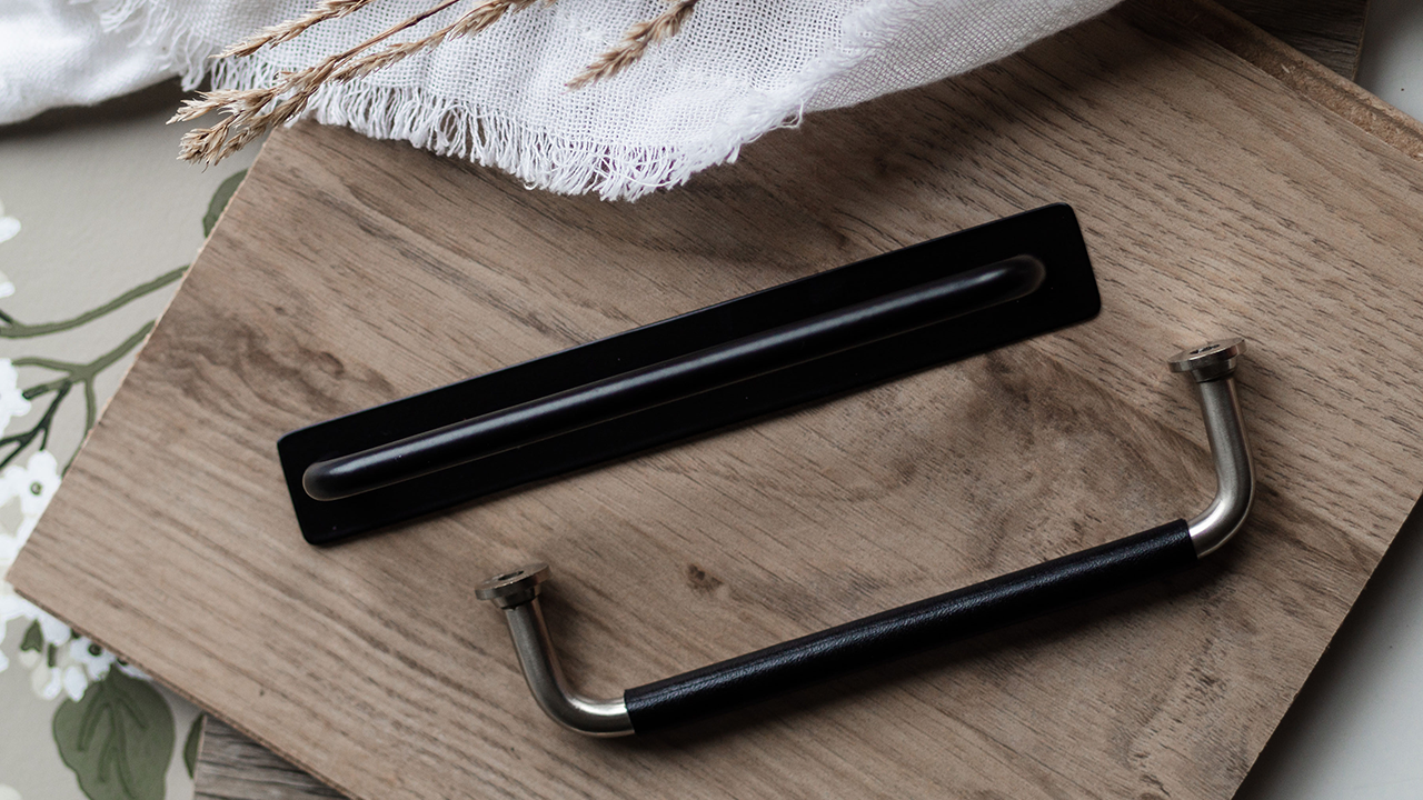 Hur döljer man märken från gamla kökshandtag?  Två svarta kökshandtag på bänkskiva i trä. Ett handtag har en svart täckbricka under sig.