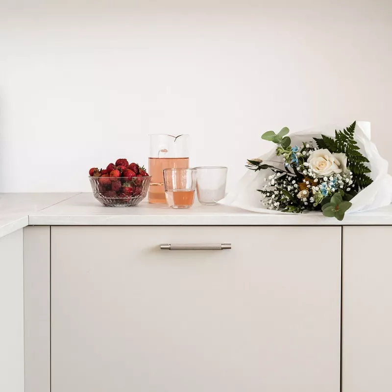 En borstad nickelhandtag monterad på en vit skåplucka med en karaff och glas med rosa dryck samt en bukett blommor ovanpå.