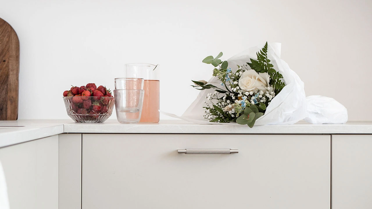 Should you have knobs or handles on kitchen drawers? En borstad nickelhandtag monterad på en vit skåplucka med en karaff och glas med rosa dryck samt en bukett blommor ovanpå.