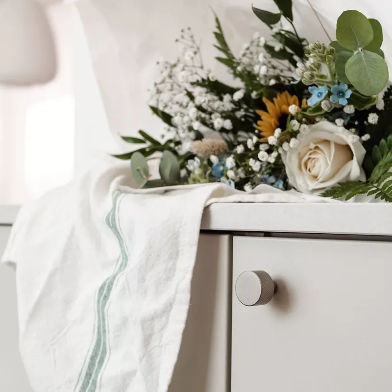 Borstad nickelknopp och handtag på en beige skåplucka, dekorerad med gröna blad och färgprover.