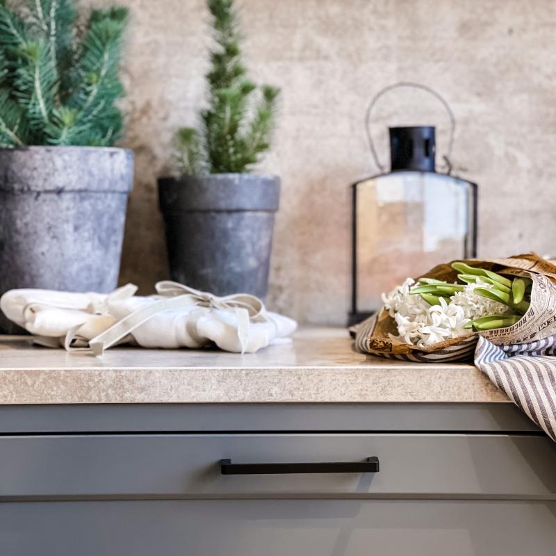 Mörkgrått kök med svarta minimalistiskt kökshandtag, mingran och hyancint på en beige bänkskiva i sten