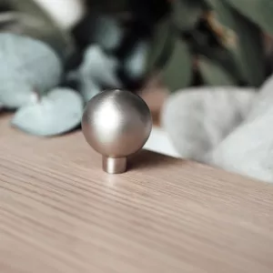 Rund knopp i rostfri look från Bona monterad på en trälucka med gröna växter och linnetyg i bakgrunden.