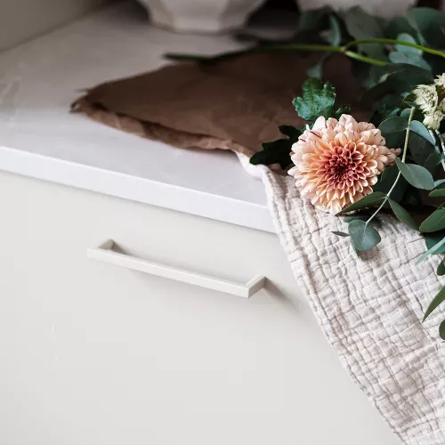 Beige handtag monterat på en vit möbel med dekorativa blommor i bakgrunden.