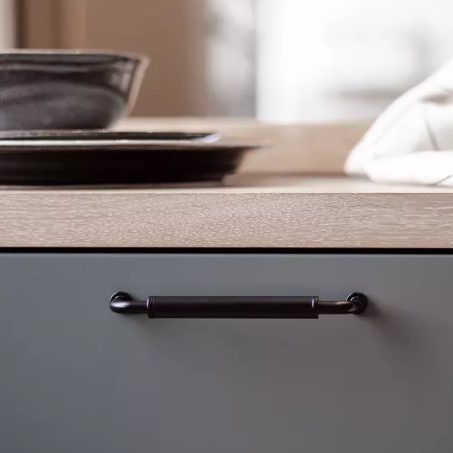 Ett kökslåds handtag i svart, i kontrast mot en ljus träbänkskiva med staplade mörka skålar ovanpå.