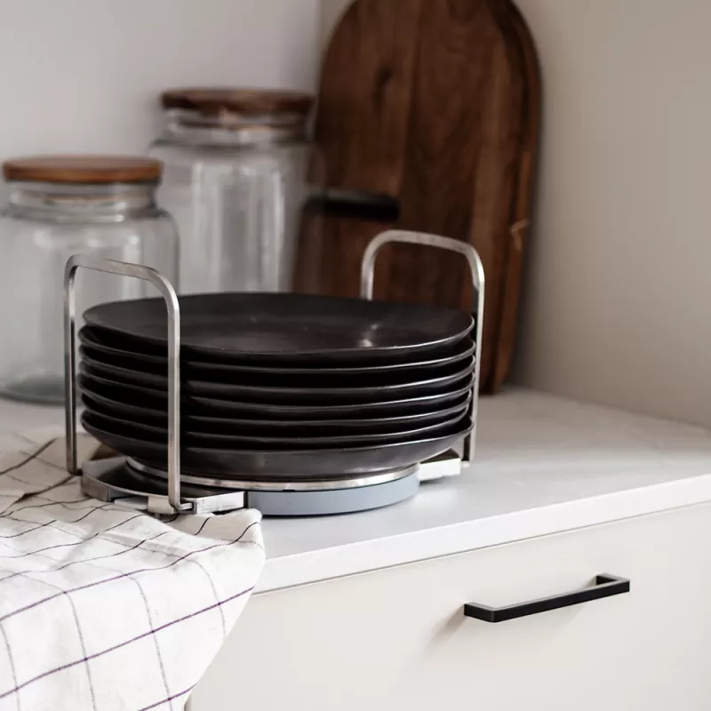En stapel svarta tallrikar i en rostfri tallrikshållare placerad på en ljus köksbänk. I bakgrunden syns glasburkar med trälock och en träskärbräda
