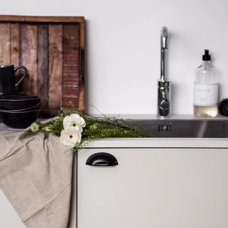 Svart skålhandtag i lantlig stil monterat på kökslucka i beige. Vårdukning med vita blommor, linneservett och träbricka framför diskho.
