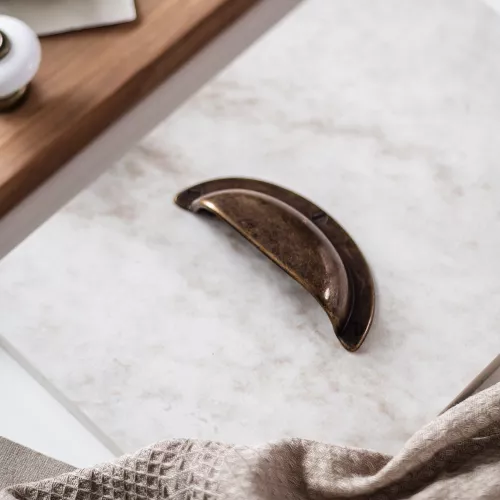 Skålhandtag i antik mässing med patinerad yta placerat på stenliknande underlag. Beige kökshandduk och porslinsknopp i bakgrunden.