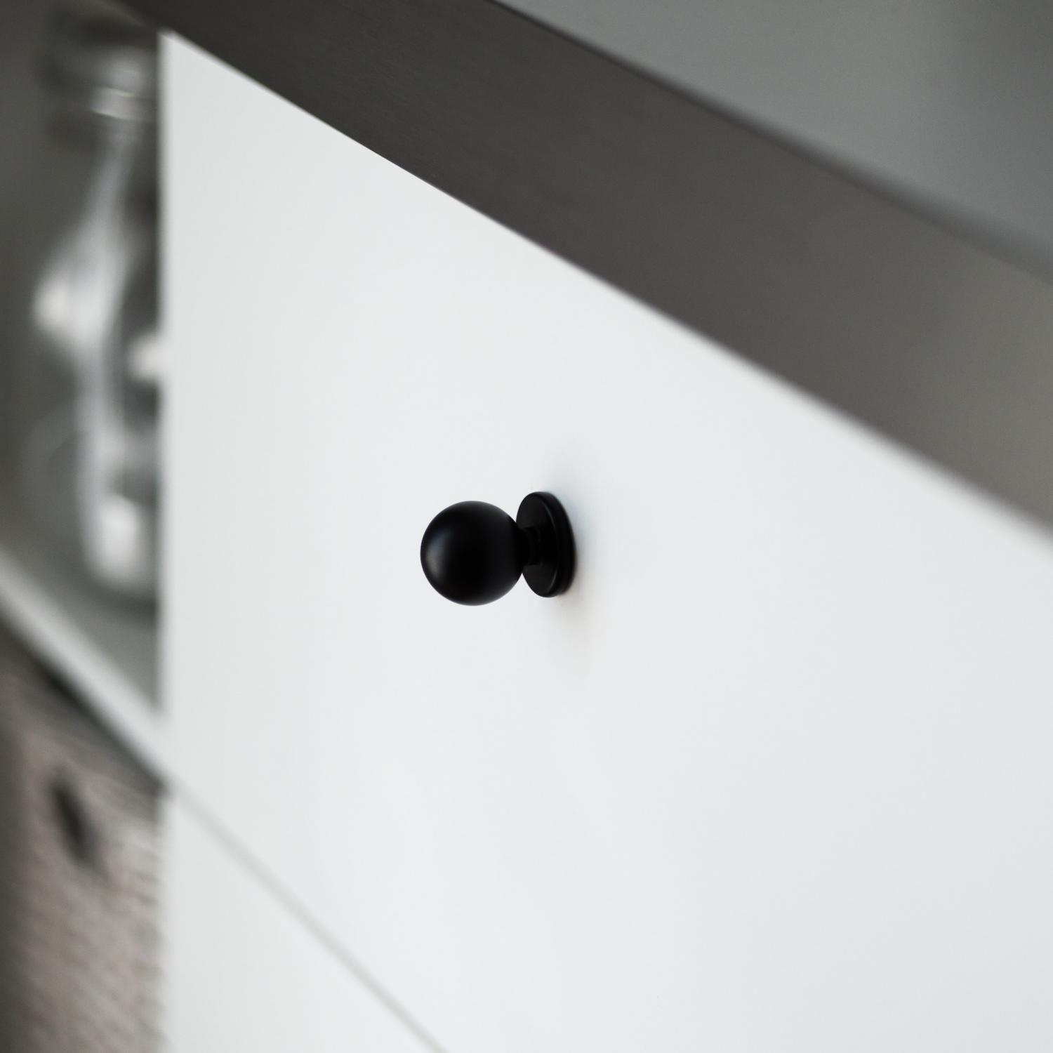 Rund svart knopp med bottenplatta, monterad på en vit skåpslucka.