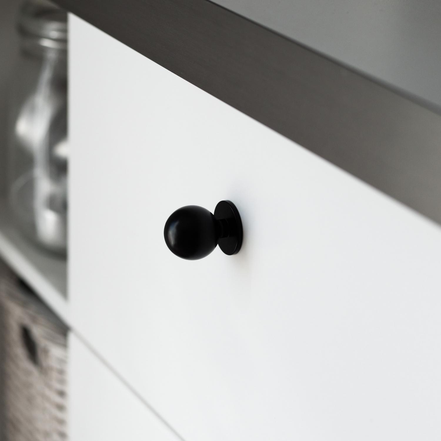 Rund svart knopp med bottenplatta, storlek 25 mm, monterad på en vit skåpslucka.