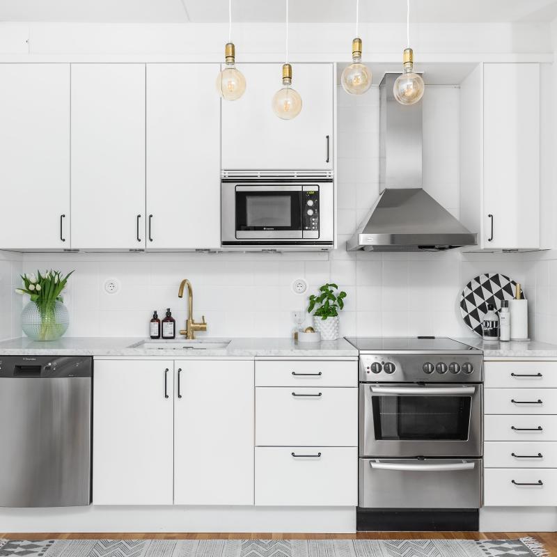 Vitt kök med mässingdetaljer och rostfria vitvaror. I taket händer en lampa i mässing och på golvet finns en mönstrad matta.