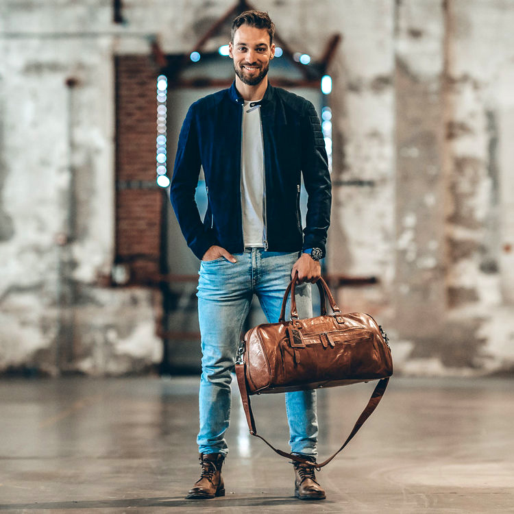 man som står framför en tegelvägg och håller en brun weekendväska i läder av modellen mainz från varumärket the  chesterfield brand