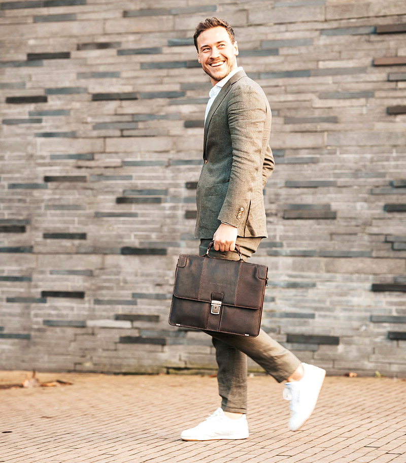 Man som går framför en grå tegelvägg bärandes på en brun läderportfölj av modellen Stuttgart tillverkad av the Chesterfield Brand