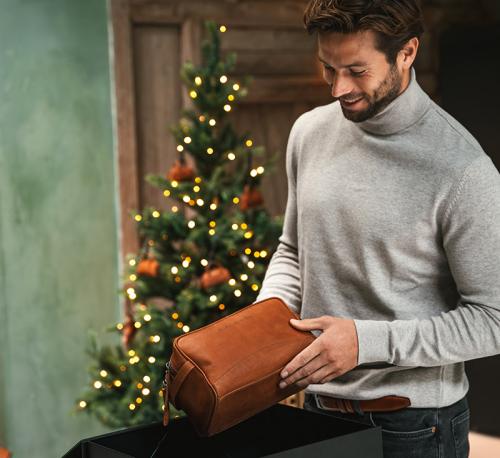 En man står framför en julgran och håller en cognacsfärgad necessär i läder