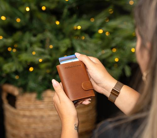 Ett par händer som håller en plånbok i cognacsfärgat läder framför en julgran