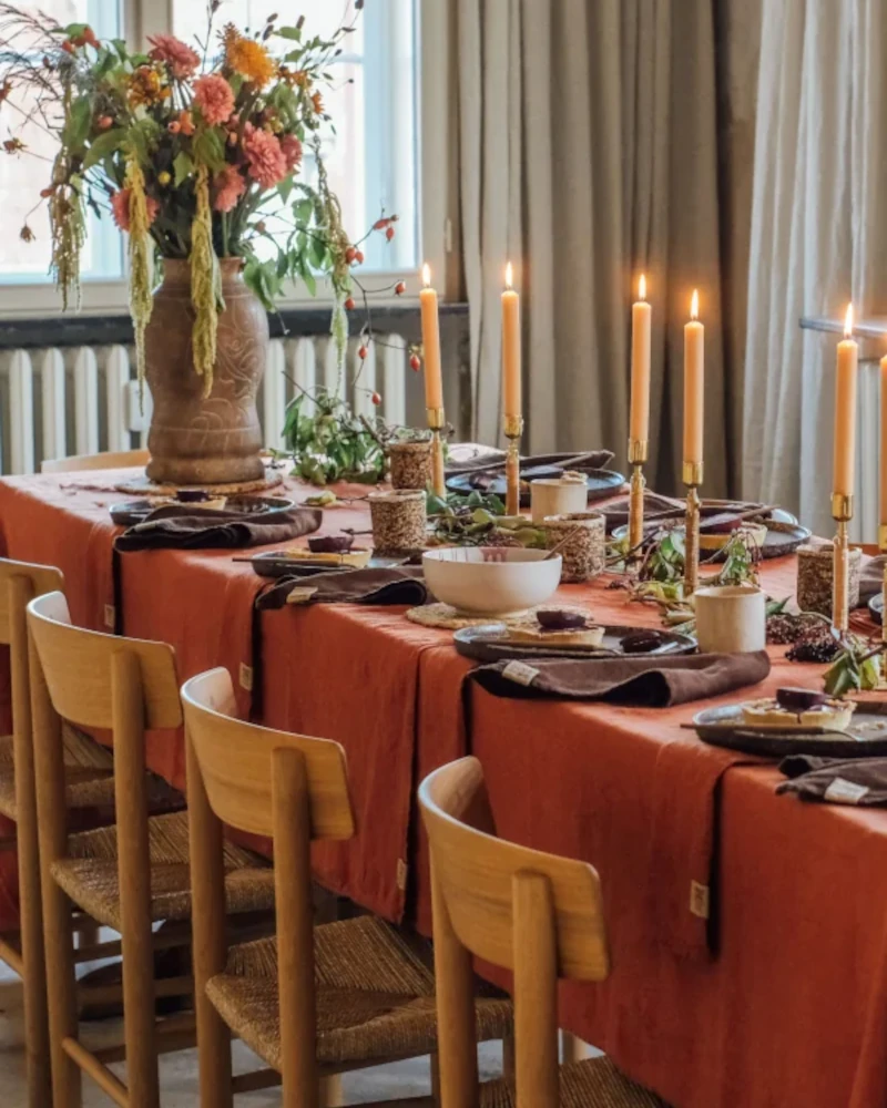 Dukat bord med en roströd linneduk i färgen Lovely Cayenne, bruna servetter, tända ljus och en stor blomsterbukett.