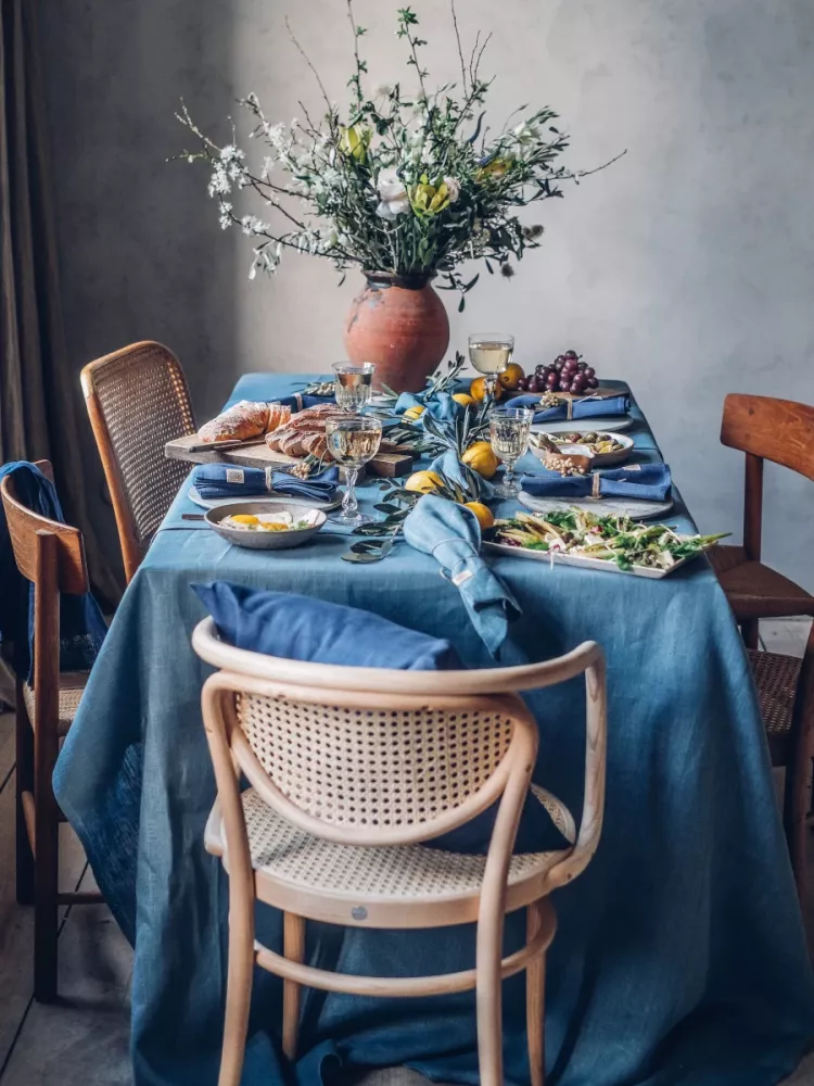 Ett matbord för en måltid med blå duk i linne, blå servetter, bröd, sallad, vindruvor och två vinglas. En stor vas med en bukett blommor fungerar som mittpunkten.
