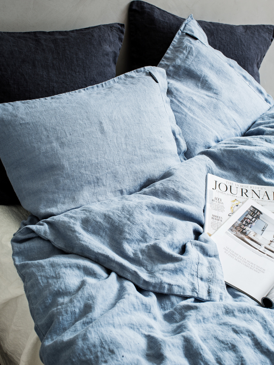 En mysig säng med rynkiga ljusblå linnesängkläder och två stora mörka kuddar. En tidning med titeln "Journal" vilar på sängen.