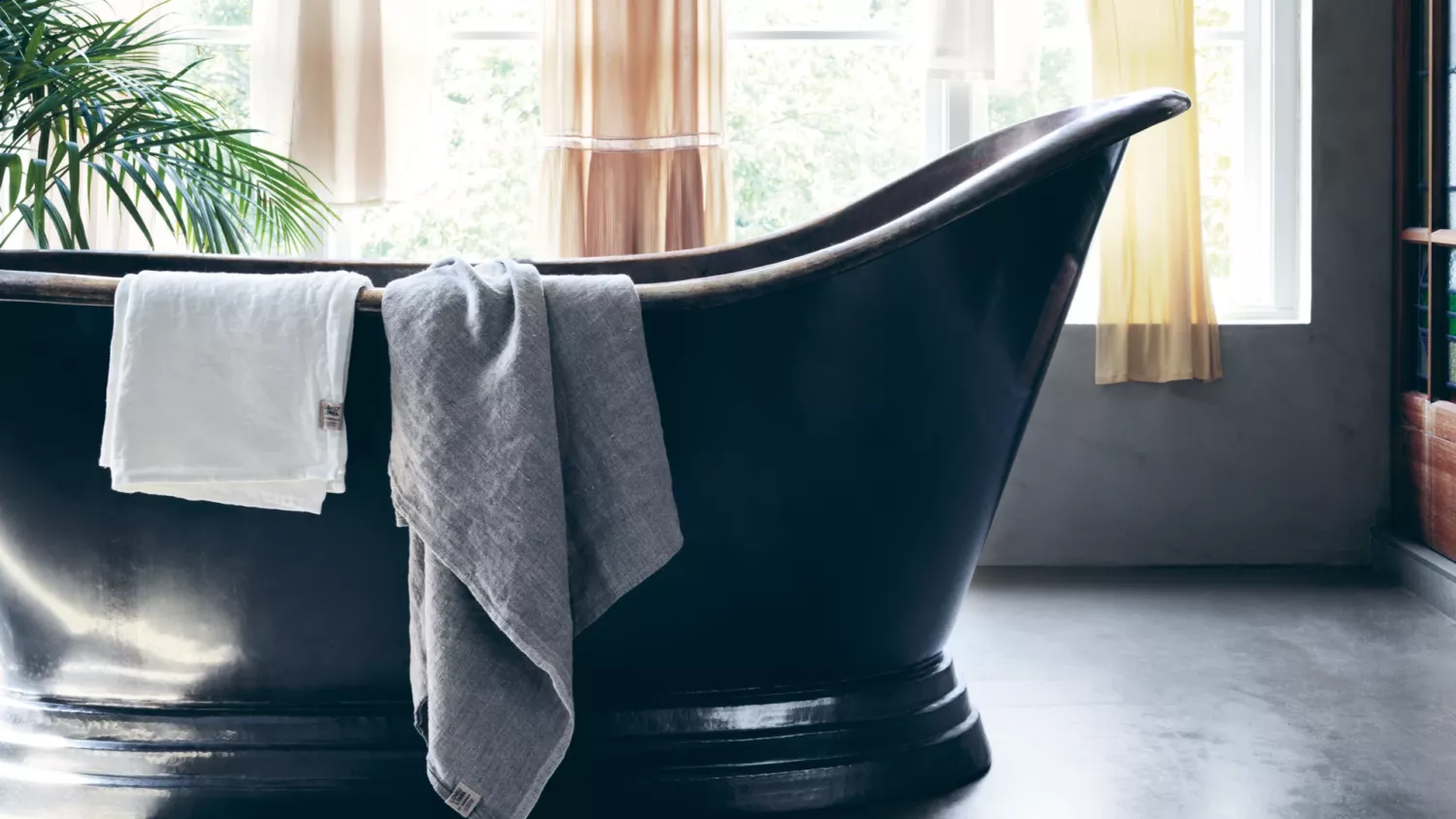 Ett svart fristående badkar med två handdukar i linne draperade över sidan, placerat i ett ljust badrum med ett stort fönster. Solljus strömmar genom linnegardiner, lyser upp utrymmet och avslöjar en krukväxt i bakgrunden.