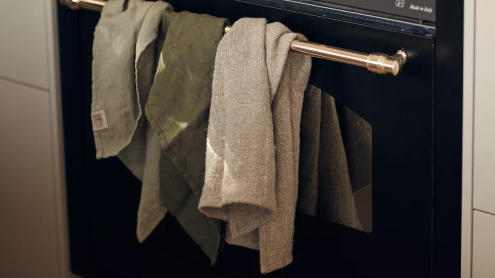 Linen in the Kitchen: Elegance and Durability Tre kökshanddukar i gröna och beige nyanser hänger över ett ugnshandtag. Ugnen är svart med ett handtag i rostfritt stål, och handdukarna ser ut att vara gjorda av linne.