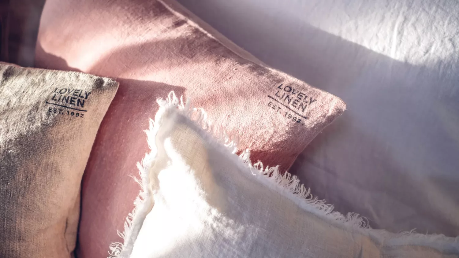 Närbild av flera kuddar i linne i olika ljusa nyanser, inklusive rosa, grädde och vitt. Texturerna sträcker sig från släta till lätt slitna kanter, vilket skapar ett arrangemang i mjuk naturlig belysning.