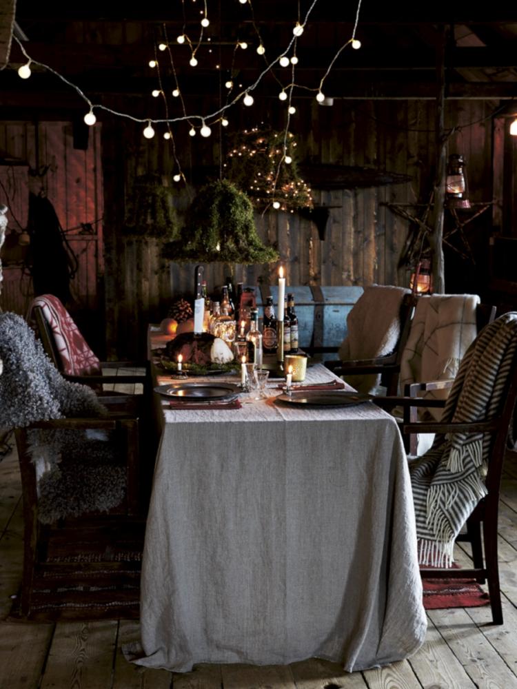 En mörk, elegant dukning med staplade blå och vita tallrikar, glas och en svart bricka med vikta servetter. Bruna, gröna glasflaskor, ljushållare med vita ljus och grönt bladverk fulländar arrangemanget.