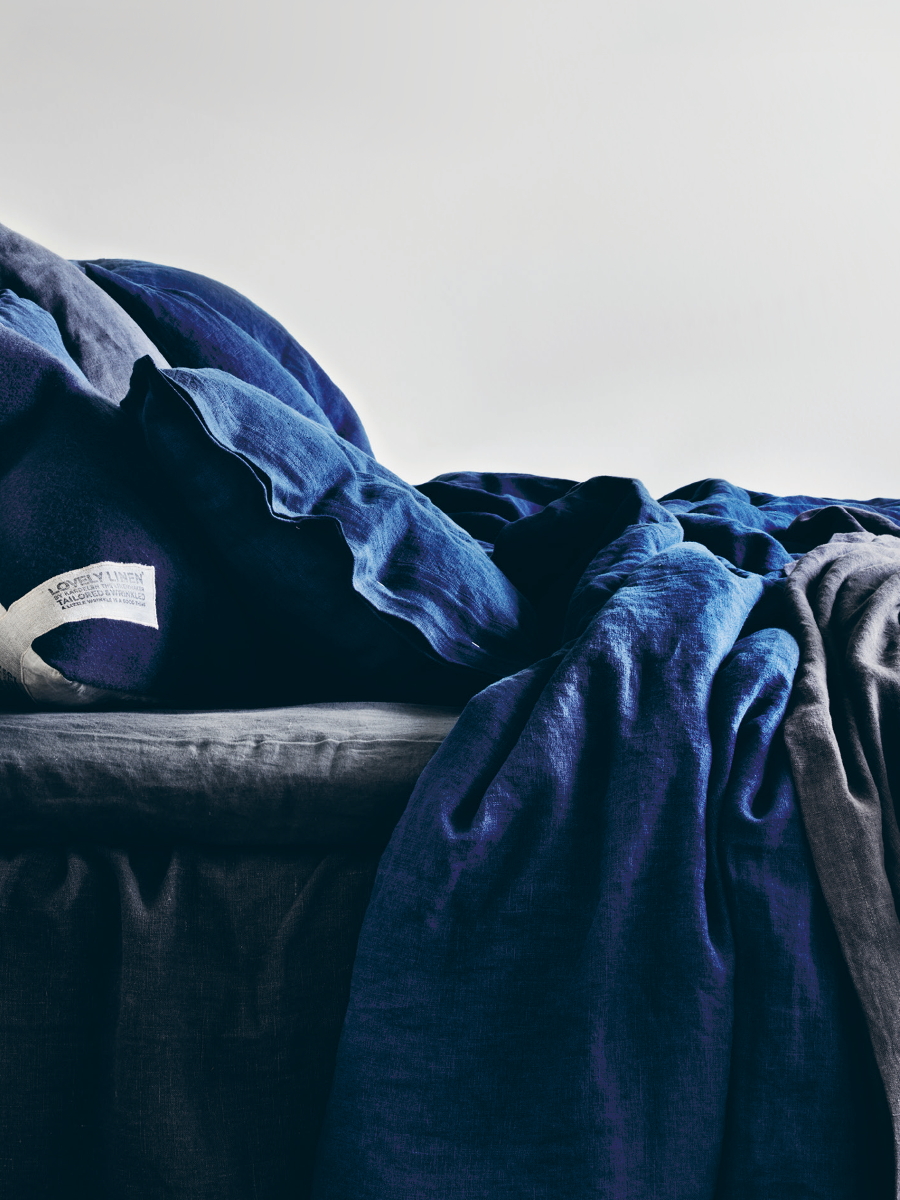 En säng med mörkblå och grå rufsiga lakan och kuddar mot en enkel bakgrund. Allt i linne.
