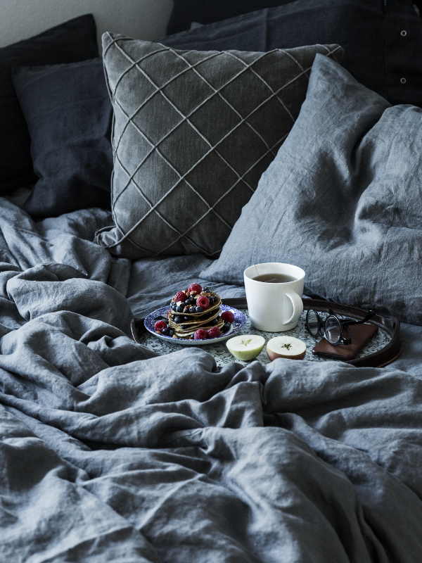 En mysig säng med gråa lakan, en bricka med en kopp kaffe, en muffins och ett litet ljus, tillsammans med dekorativa kuddar i bakgrunden.