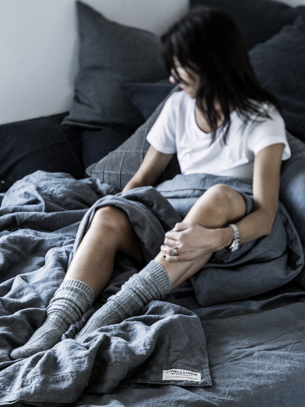 Person som sitter på en säng, klädd i en vit t-shirt och gråa strumpor, med en grå filt som täcker sina ben.