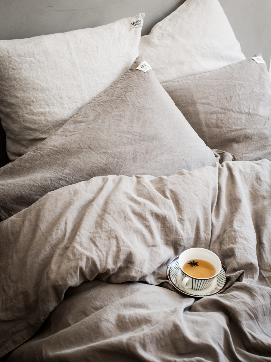 Obäddad säng med beige sängkläder i linne. En kopp kaffe och en liten tårta står på ett fat placerat på sängen. Naturligt ljus lyser upp scenen.