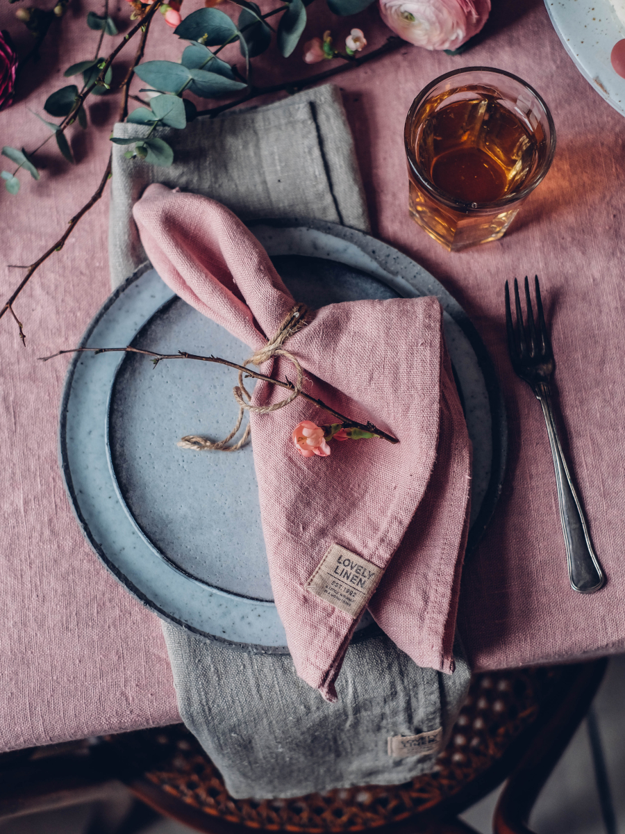 En rustik dukning har en grå tallrik med en rosa linneservett bunden med garn och en liten blomma. Ett glas bärnstensfärgad vätska och en gaffel finns bredvid. Bordet är täckt med en rosa duk, och det finns kvistar av grönska och blommor.
