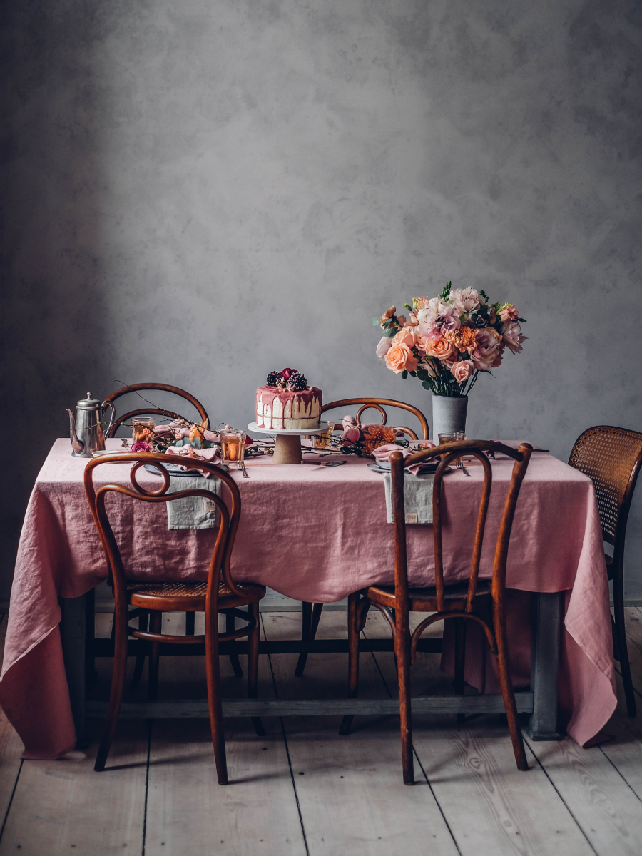 Ett matbord med en rosa duk, trästolar och en tårta prydd med rosa glasyr och bär. En stor bukett blommor i rosa och persika toner sitter i ena änden av bordet.
