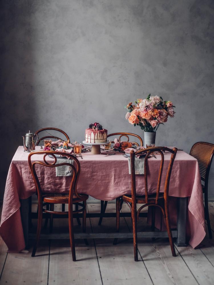 Ett matbord med en rosa duk, trästolar och en tårta prydd med rosa glasyr och bär. En stor bukett blommor i rosa och persika toner sitter i ena änden av bordet.