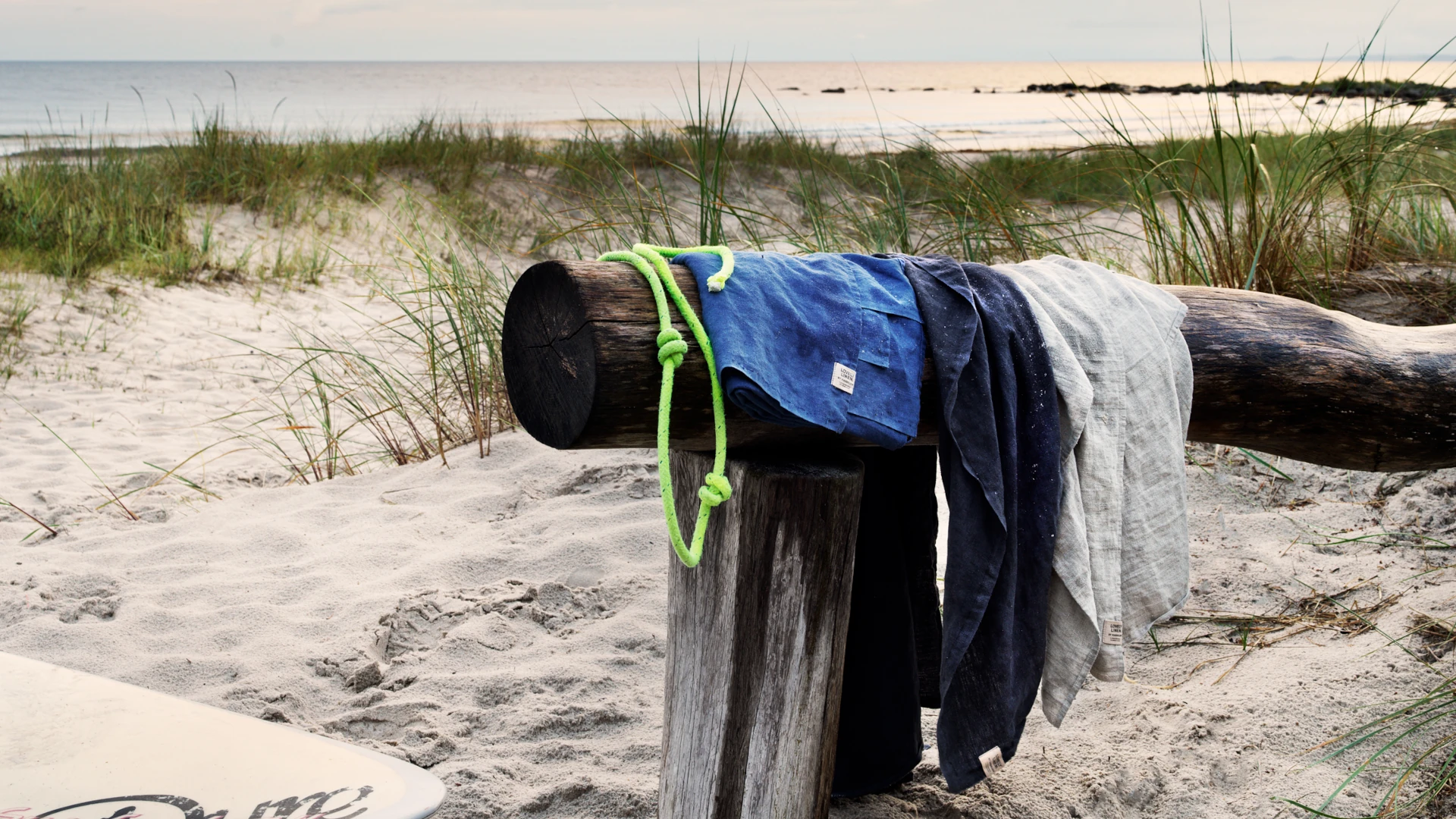 Baddukar i linne hönger över en stock på en sandstrand med gräs och en fridfull havsutsikt i bakgrunden.